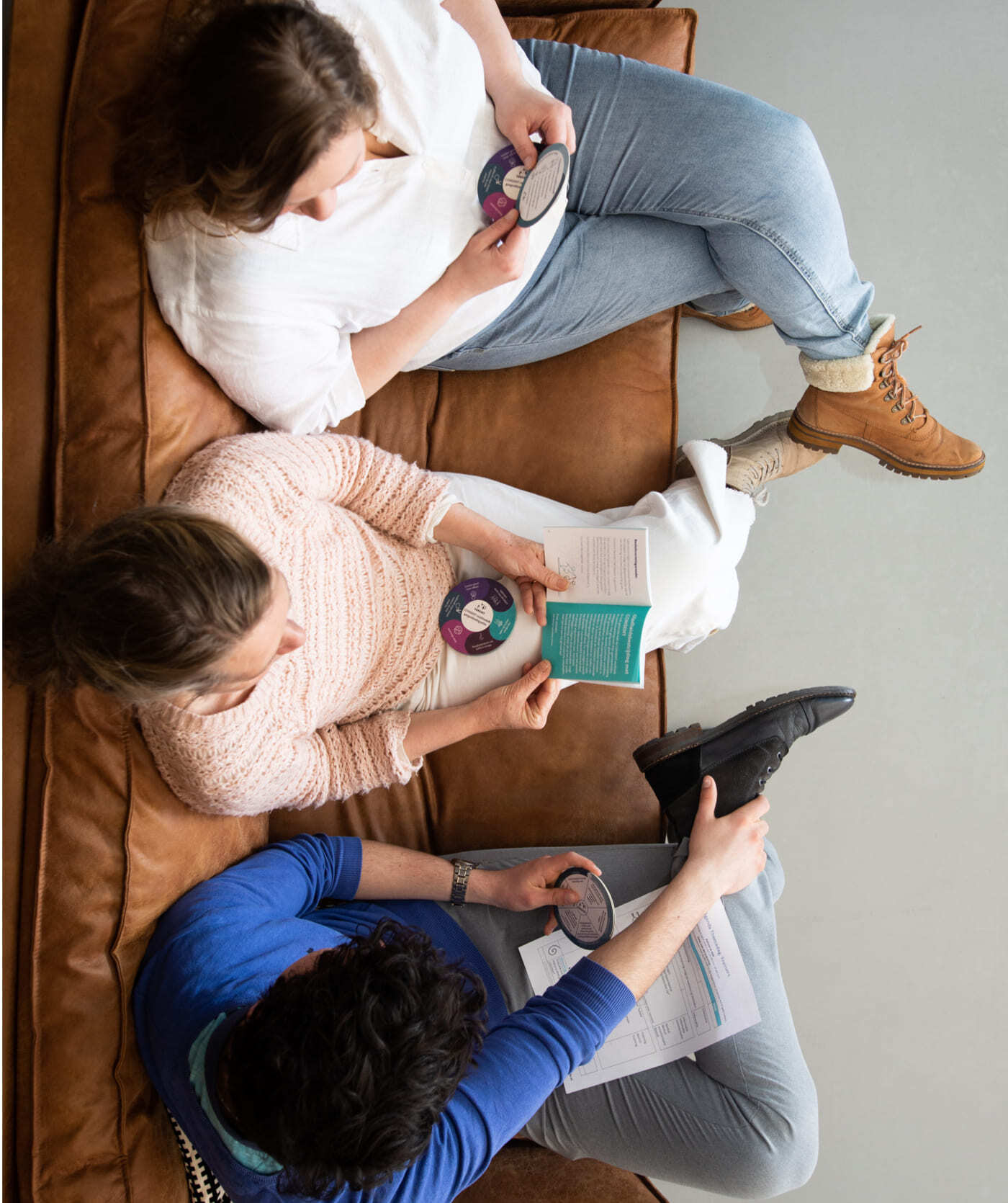 Drie mensen zitten op een bruine bank, houden ronde kaarten vast en lezen boekjes. Mogelijk zijn ze bezig websites of online diensten te bekijken, van bovenaf gezien.