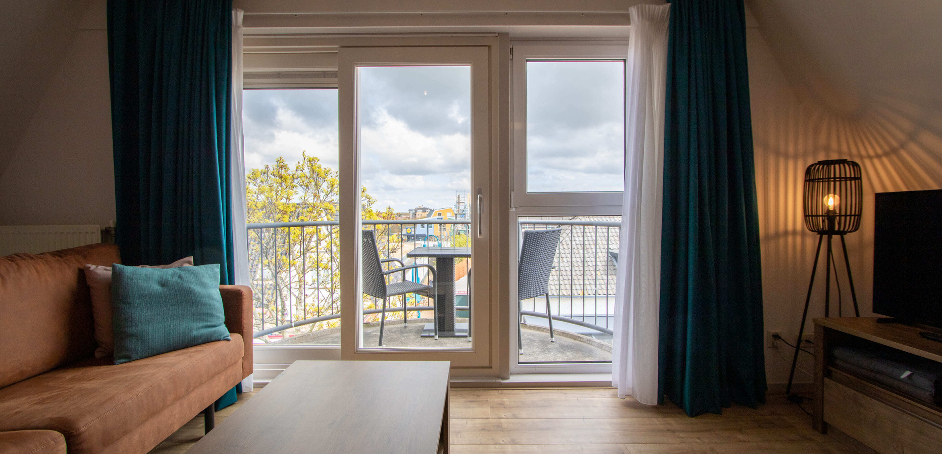 Woonkamer met bruine bank, blauwgroen kussen, salontafel en staande lamp, met uitzicht op een glazen deur naar een klein balkon met twee stoelen en uitzicht op de stad – perfecte inspiratie voor webshops of websites die online diensten aanbieden.