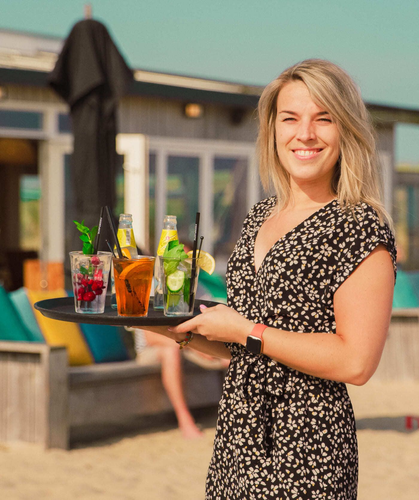 Een vrouw in een jurk met een print staat op een zandstrand met een dienblad met drie cocktails en flesjes drinken, met een gebouw aan het strand op de achtergrond. De perfecte inspiratie voor websites of webshops in de horeca.