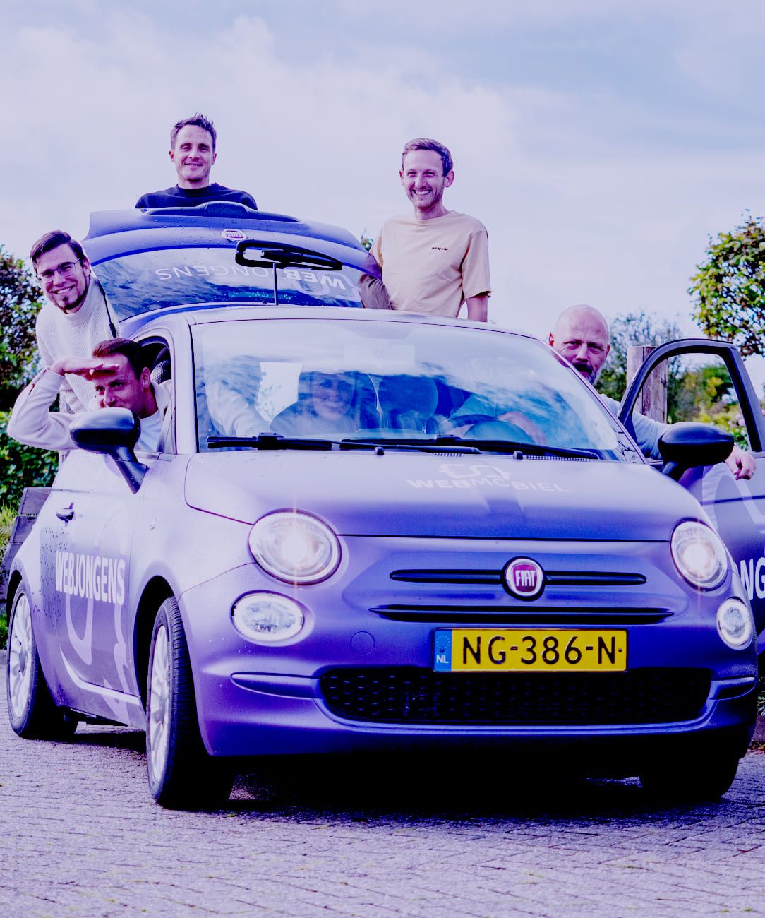 Vijf mannen poseren bij een paarse Fiat 500 met het Nederlandse kenteken NG-386-N; twee zitten in de auto, drie staan erbuiten en één houdt de autodeur open – wellicht om te praten over websites of online diensten.