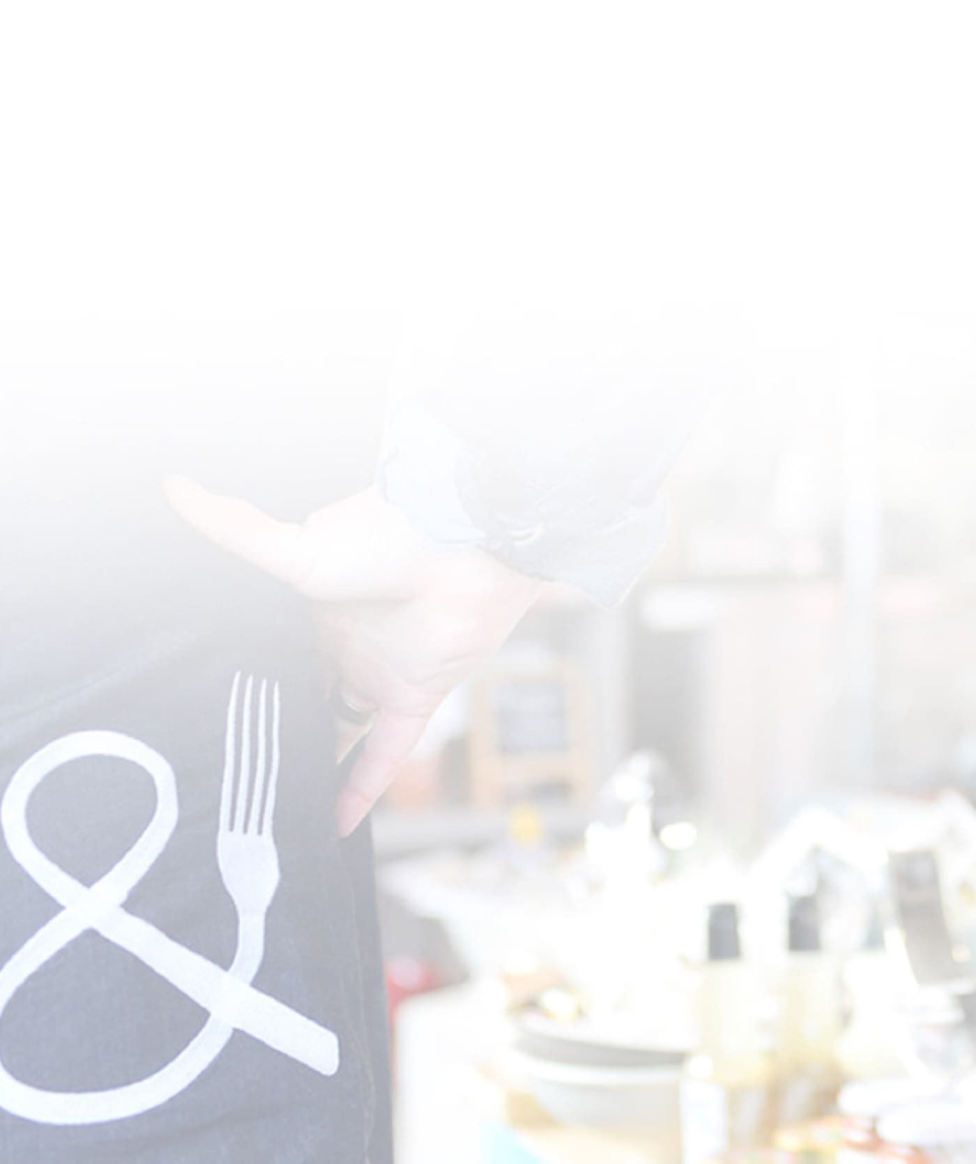 Een persoon in een donkere broek met een wit ampersand en een vork staat in een vage, goed verlichte binnenomgeving, mogelijk gerelateerd aan webshops of online diensten, met flessen en verschillende artikelen op een tafel.